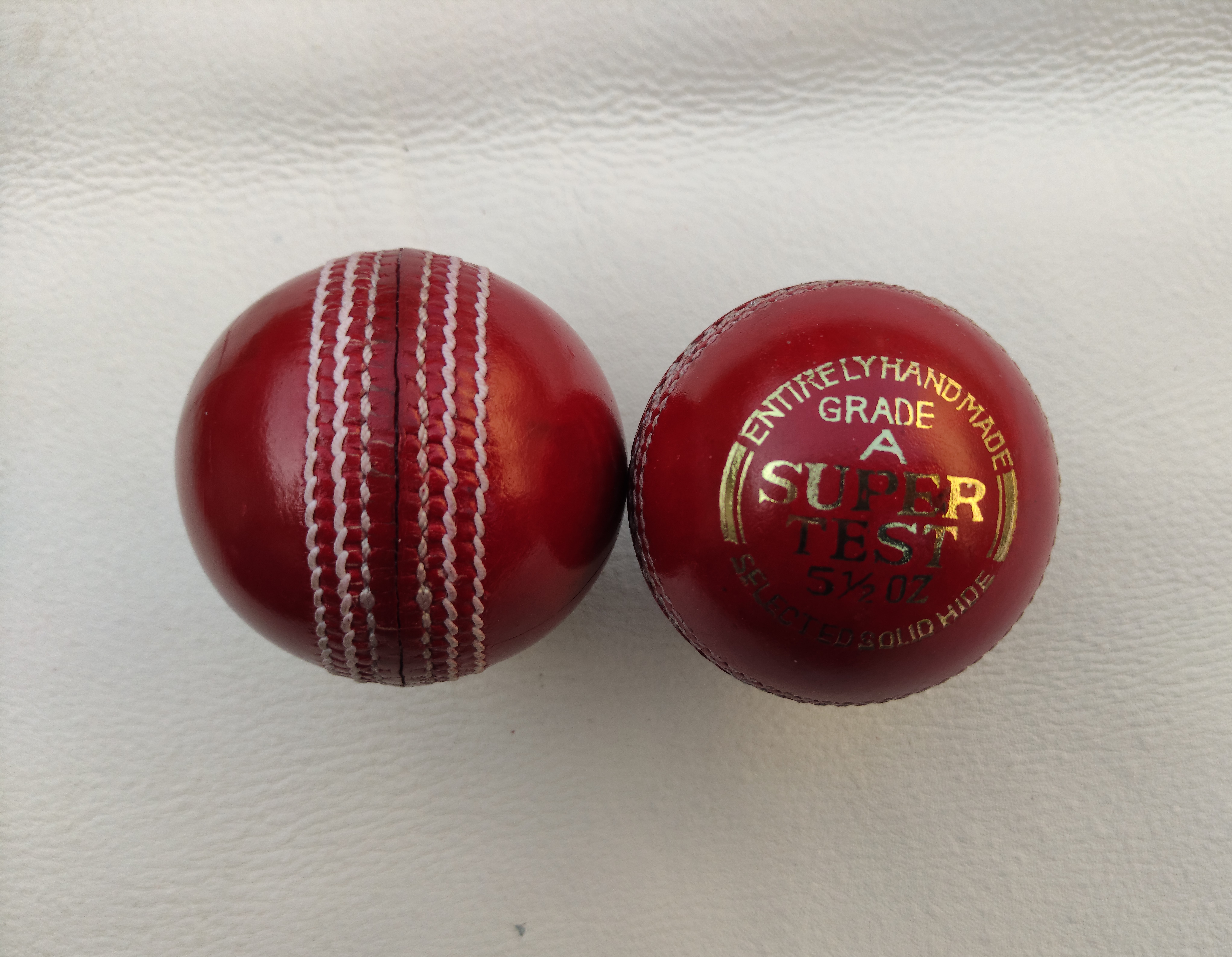Two red 4-piece standard seams leather cricket balls, one shown from the front and the other from the side angle, placed clearly to display seam stitching and leather texture.