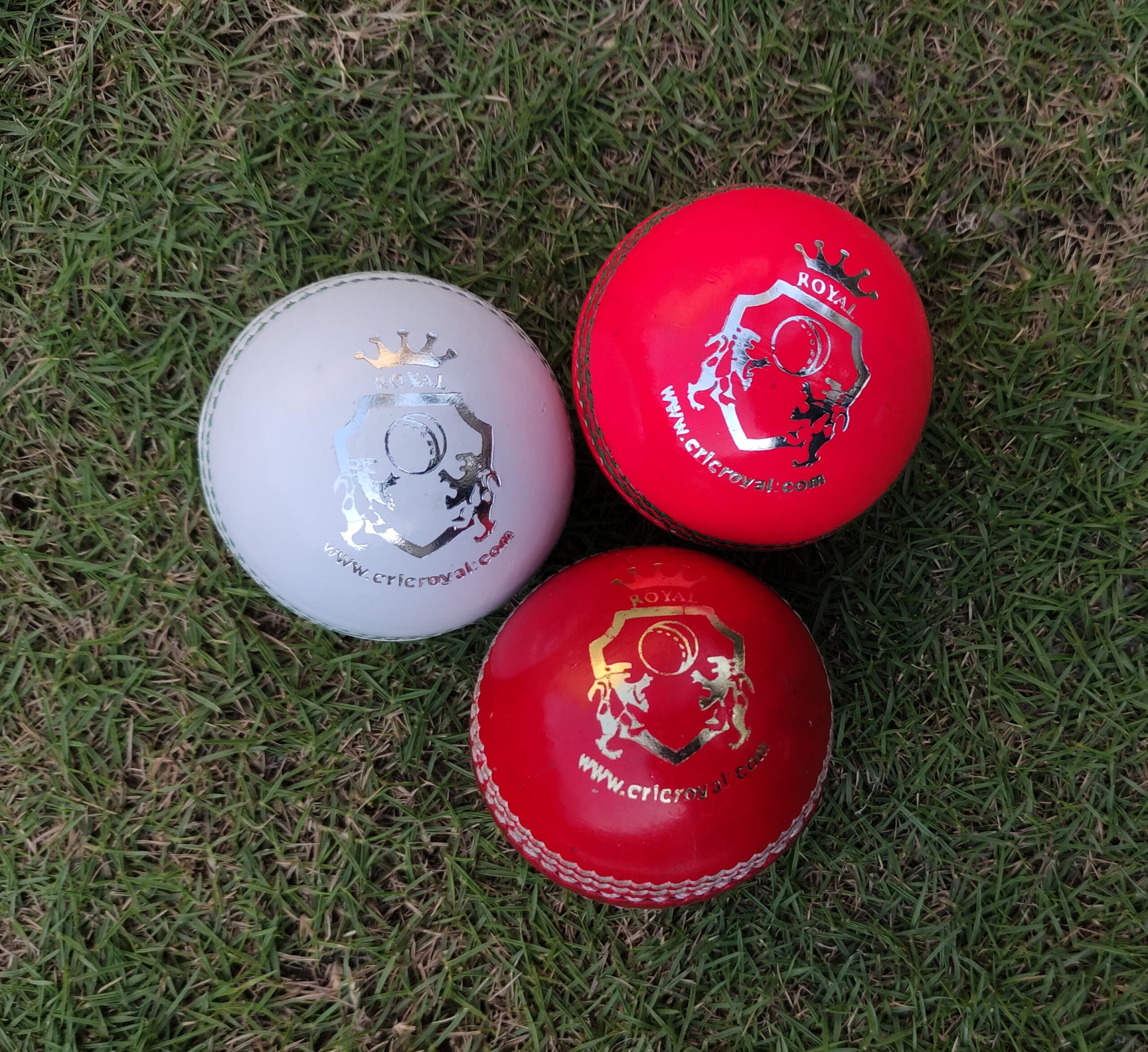 Three Kids Leather Cricket Hard Balls, each in a different color (red, white, and pink), placed on green grass in front-facing view, showcasing variety for junior training and play.