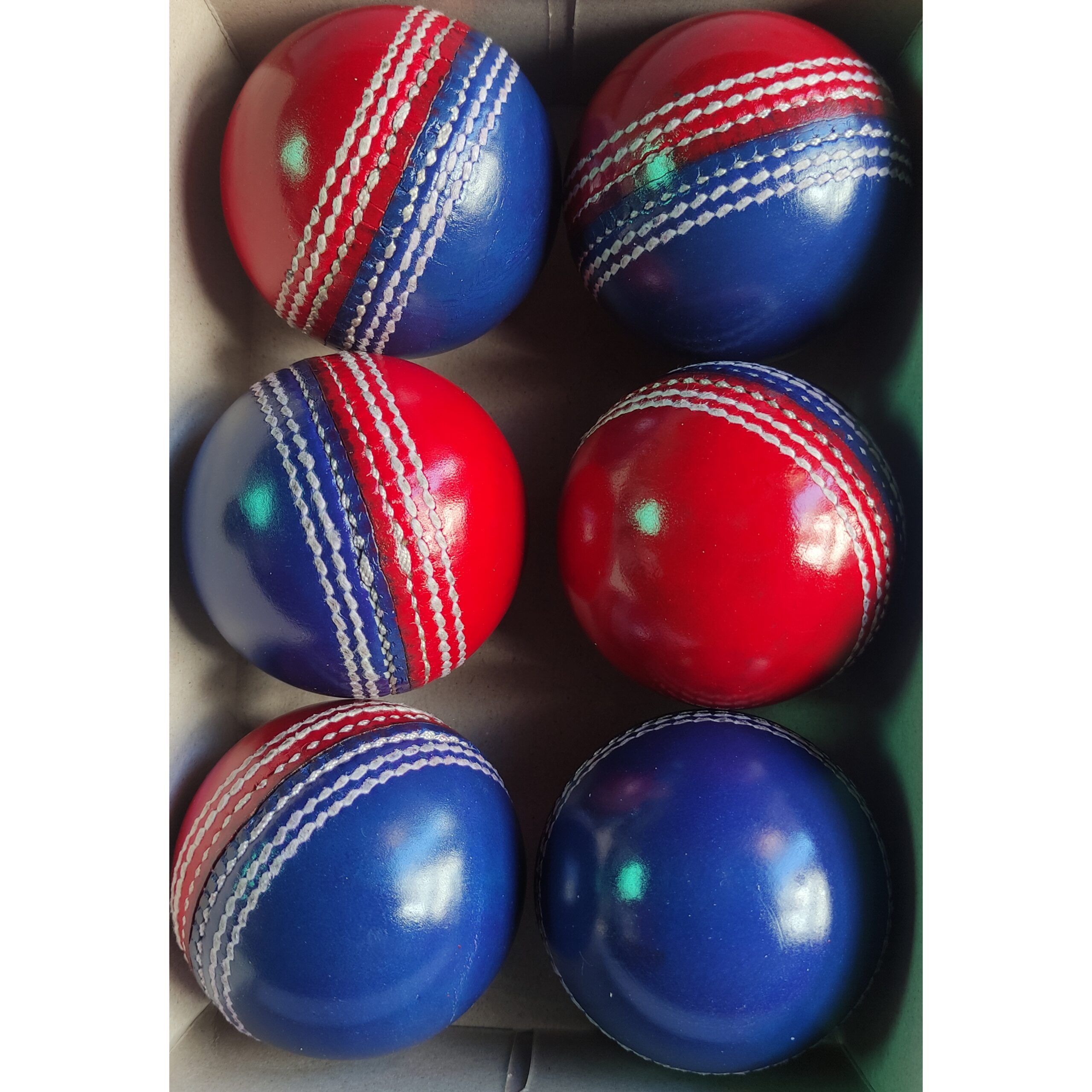 Six identical red and dark blue dual color leather cricket balls placed inside a box, captured from different angles to show side and front views, perfect for training and coaching use.